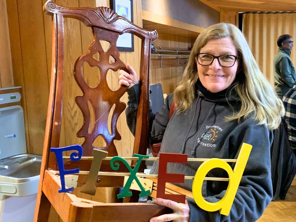 Women smiling holding wooden chair and sign that says, 'fixed'.